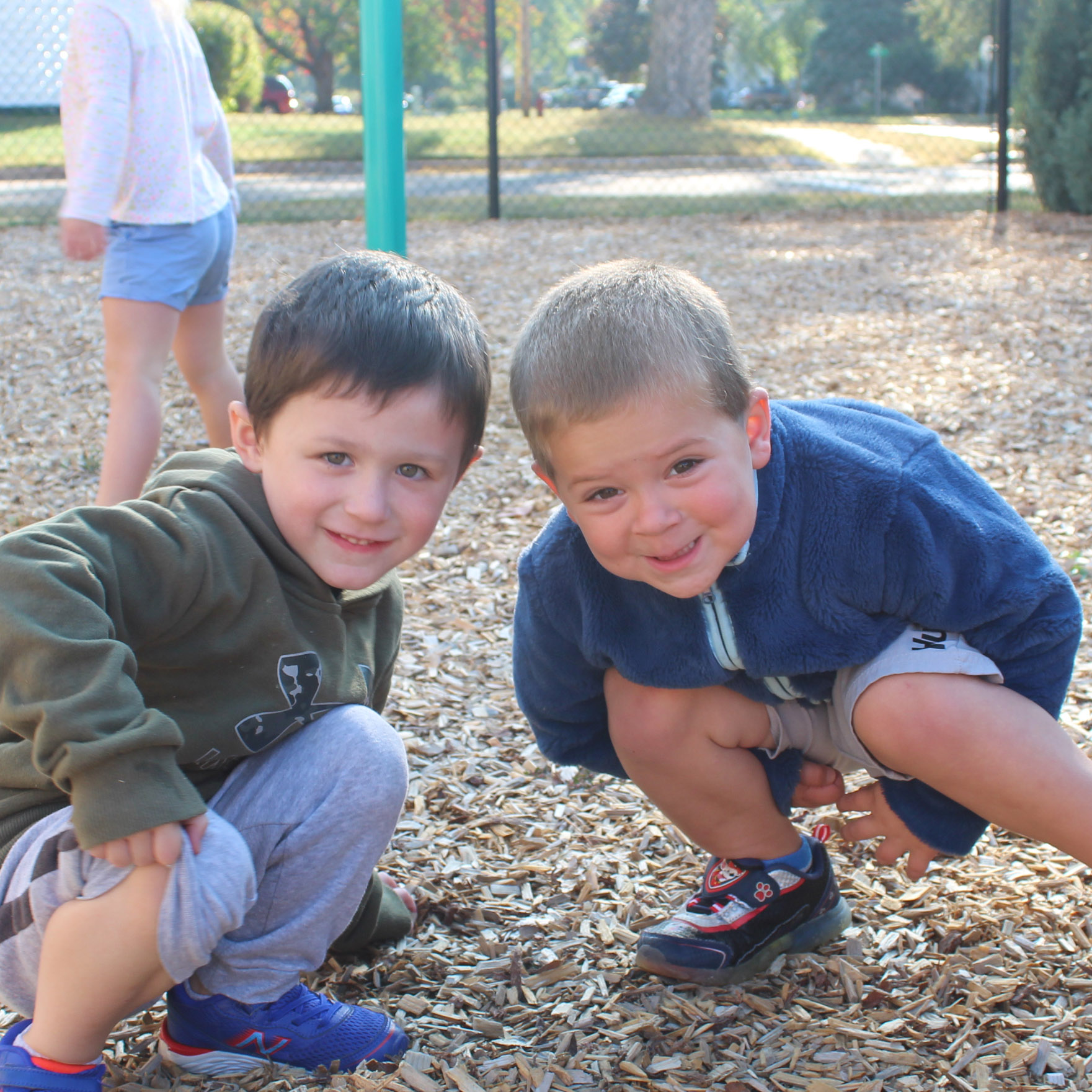 Two children crouch down and smile for a photo while at a playground